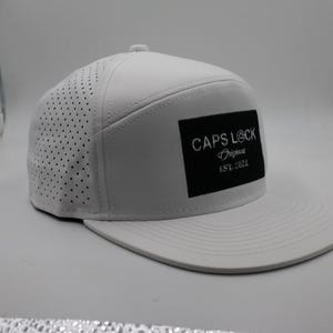 May include: A white baseball cap with a flat brim. The cap features a perforated side panel and a black rectangular patch with the text "CAPS LOCK Original EST. 2022".