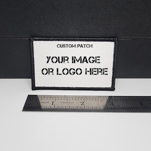 以下が含まれることがあります： 「CUSTOM PATCH」、「YOUR IMAGE」、「OR LOGO HERE」と黒字で書かれた白い長方形のカスタムパッチ。黒い縁取り。ステンレス製の定規が下にあり、センチメートル単位の測定値が表示されています。