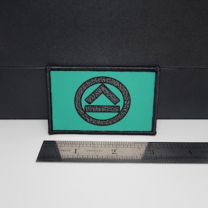 Puede incluir: Parche rectangular con fondo turquesa y borde negro. El parche presenta un diseño circular gris con un símbolo de flecha estilizado. Una regla de acero inoxidable se coloca debajo.
