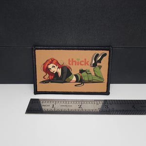 May include: A rectangular patch featuring a cartoon illustration of a woman with red hair, wearing a black top, green trousers, and black shoes. The word "thick" is printed on the patch. The patch is approximately 7.6 cm long.