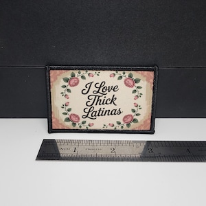 May include: Rectangular patch with the text "I Love Thick Latinas" surrounded by a floral wreath. The patch has a black border and a cream-colored background with pink roses. A ruler is placed below the patch.