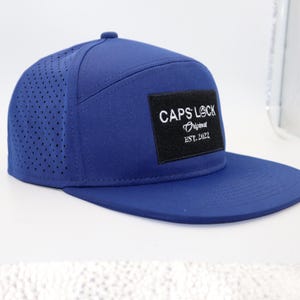 May include: A blue baseball cap with a flat brim. The cap features a perforated side panel and a rectangular patch with the text "CAPS LOCK Original EST. 2022".