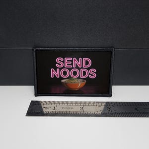 May include: A rectangular patch with the words "SEND NOODS" in pink neon lettering above a bowl of noodles. The patch is black with a black border. A ruler is below the patch.