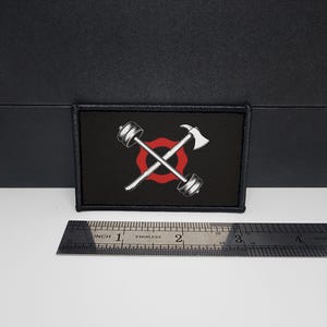 May include: Black rectangular patch featuring a white barbell and axe crossed over a red circle on a black background. A stainless steel ruler with inch measurements is below the patch.
