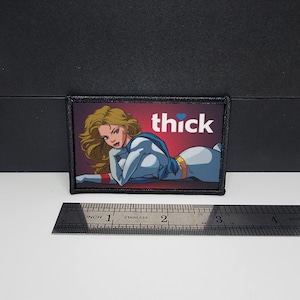 May include: Rectangular patch with a cartoon illustration of a woman in a blue and white outfit, and the word "thick" in white text. The patch is bordered in black and is approximately 7.6 cm wide.