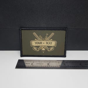 Puede incluir: Parche rectangular verde oliva con borde negro. El parche presenta un diseño temático de golf con palos de golf cruzados, una pelota de golf y el texto "YOUR • TEXT" en una pancarta. El parche mide aproximadamente 7,6 cm de largo.