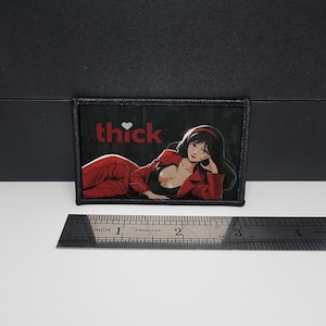 May include: A rectangular patch with a black border. The patch features the word "thick" in red with a heart symbol, and an illustration of a woman in a red suit and black top. A ruler is placed below the patch.