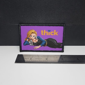 May include: A rectangular patch with a purple background featuring an anime-style illustration of a woman with blonde hair and the word "thick" in yellow. The patch is bordered with black stitching and includes a ruler for scale.