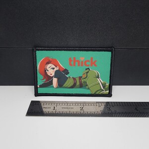 May include: A rectangular patch with a cartoon illustration of a woman with red hair and green clothing, lying down. The word "thick" is written in red letters above the woman. The patch is approximately 3 inches wide.
