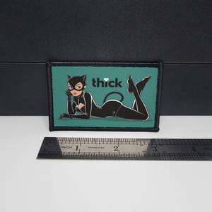 May include: A rectangular patch with a cartoon illustration of a woman in a black cat suit, mask, and gloves, with the word "thick" and a heart symbol. The background is teal, and the patch is outlined in black. A ruler is below the patch.