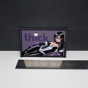 Peut inclure: Écusson rectangulaire avec un fond violet portant le mot "thick" avec un symbole de cœur et une illustration d'une femme en costume de super-héros noir et blanc. Une règle est placée en dessous.