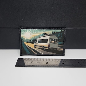 May include: A rectangular patch featuring a white camper van driving on a road towards a bright sun. The background includes mountains and trees. The patch has a black border and is approximately 3 inches wide.
