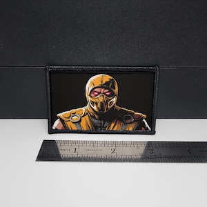 May include: Rectangular patch featuring a character in a yellow and black outfit with red eyes, set against a black background. The patch is approximately 3 inches wide. A stainless steel ruler is placed below the patch.