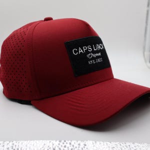 May include: A burgundy baseball cap with a curved brim. The cap features a perforated side panel and a black square patch with white text that reads "CAPS LOCK Original EST. 2022".