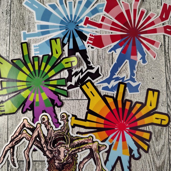 May include: A collection of four die-cut stickers featuring stylized figures with radiating sunburst patterns and the text THE THING, alongside one detailed sticker of a multi-legged creature with prominent eyes and a textured, hairy body.