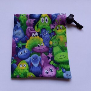 May include: A small, square drawstring bag with a vibrant pattern of cartoon monsters in green, purple, blue, and yellow. The bag has a black drawstring closure, likely made of fabric.