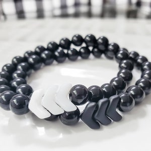 May include: Two beaded bracelets. The bracelets are made of black, round beads. One bracelet has white arrow-shaped accents, and the other has black arrow-shaped accents. The bracelets are displayed on a white surface.
