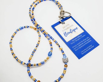 May include: A beaded lanyard featuring alternating blue, gold, and silver seed beads with a silver-toned metal chevron charm and a lobster clasp. Attached is a blue rectangular tag with white text reading Down Syndrome Boutique and Manda Gifford.