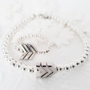 May include: A silver bracelet and ring set with a chevron arrow design. The bracelet is made of small silver beads and the ring is a simple band with a chevron arrow charm.