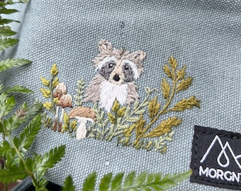 May include: A light blue fanny pack with a black zipper and embroidered raccoon design. The raccoon is surrounded by embroidered plants and mushrooms. The bag has a black label with the word MORGNTAL.