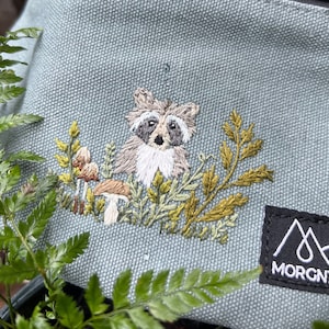 Puede incluir: Riñonera azul claro con cremallera negra y diseño de mapache bordado. El mapache está rodeado de plantas y setas bordadas. La bolsa tiene una etiqueta negra con la palabra MORGNTAL.