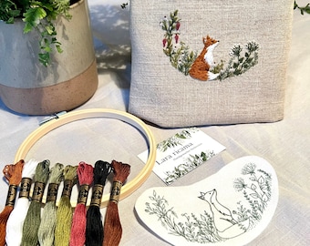 May include: A beige linen pouch with an embroidered fox design, surrounded by a floral wreath. The image also shows embroidery floss in various colors, an embroidery hoop, a needle, and a small card with the text "Lara ricama".