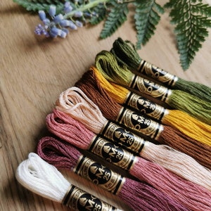 Könnte beinhalten: Eine Auswahl an DMC Stickgarn in verschiedenen Farben, darunter Weiß, Rosa, Braun und Grün. Jede Strähne ist mit dem DMC-Logo und Produktinformationen versehen. Das Garn liegt auf einer Holzoberfläche, mit einem Zweig im Hintergrund.