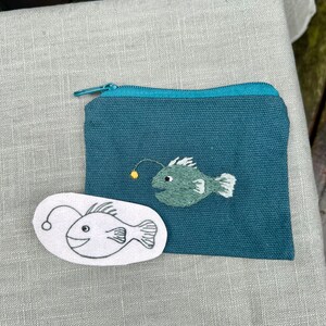 May include: A teal zippered pouch with a white outline of a anglerfish and a green embroidered anglerfish.