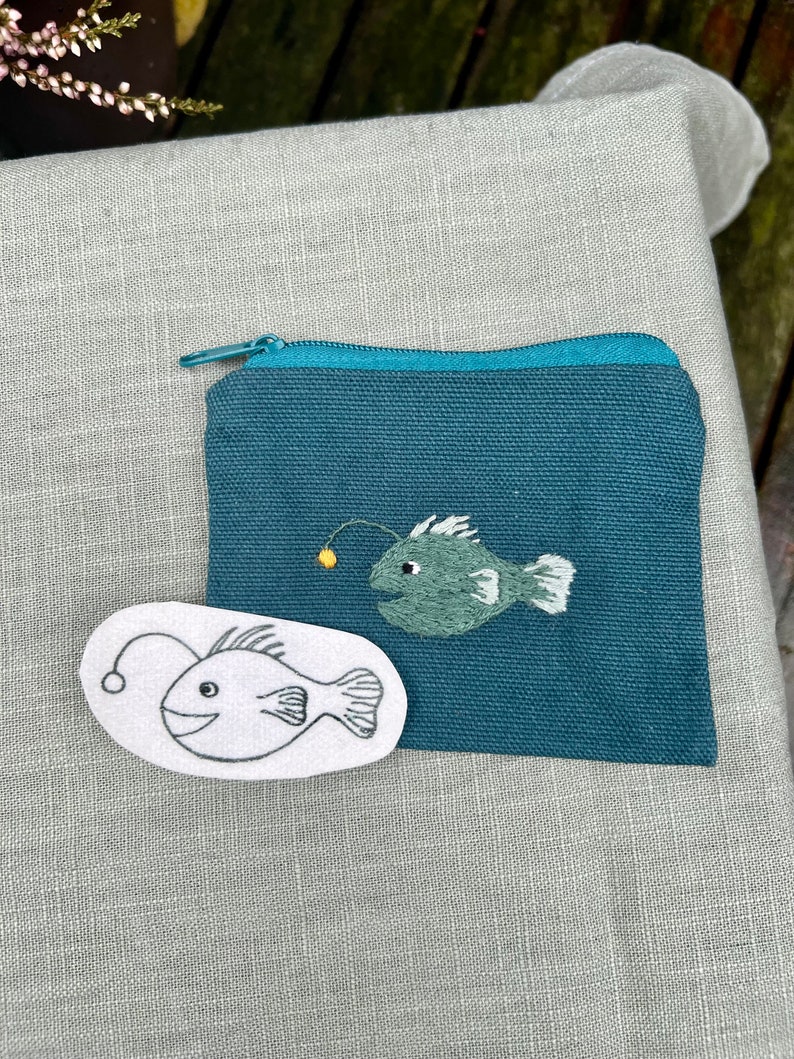 May include: A teal zippered pouch with a white outline of a anglerfish and a green embroidered anglerfish.