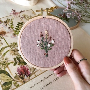 Könnte beinhalten: Ein rosa Stoff-Stickrahmen mit einem Blumenmuster aus weißen Blüten und grünen Blättern. Der Rahmen ist aus Holz und hat einen goldenen Metallring.
