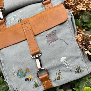 May include: A light blue canvas backpack with brown leather straps and a flap. The backpack is decorated with embroidered sea creatures, including a stingray, jellyfish, and fish.