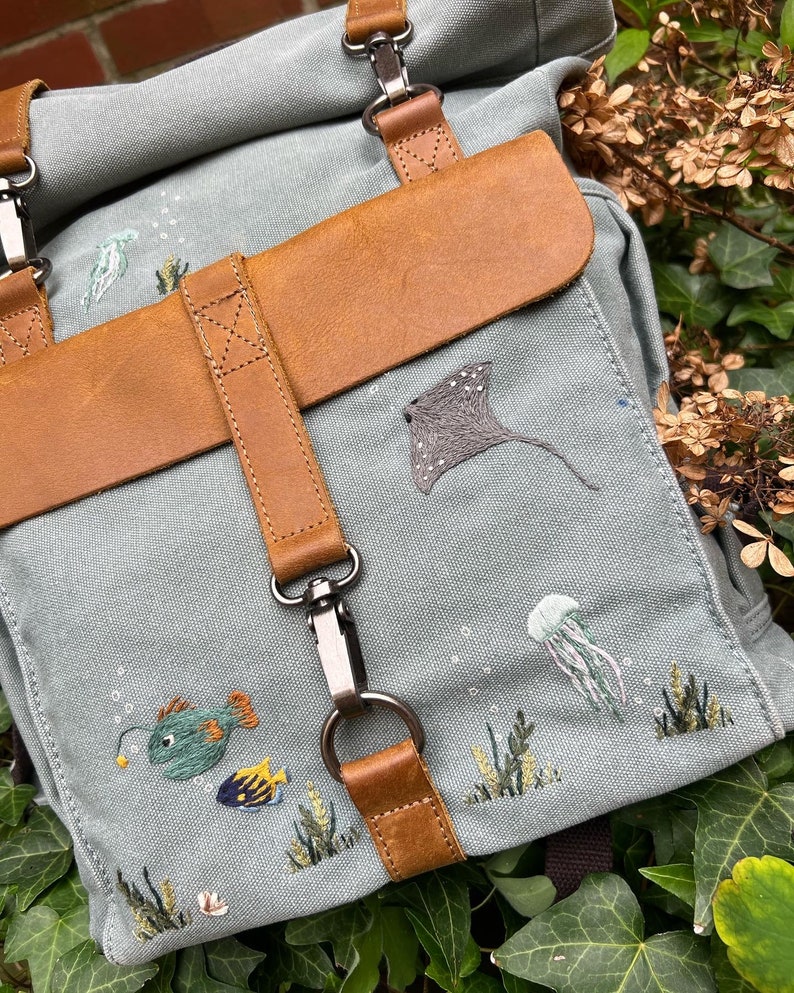 May include: A light blue canvas backpack with brown leather straps and a flap. The backpack is decorated with embroidered sea creatures, including a stingray, jellyfish, and fish.