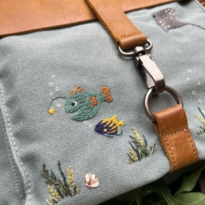 May include: A light blue canvas backpack with a brown leather strap and buckle. The backpack features embroidered images of fish and seaweed.