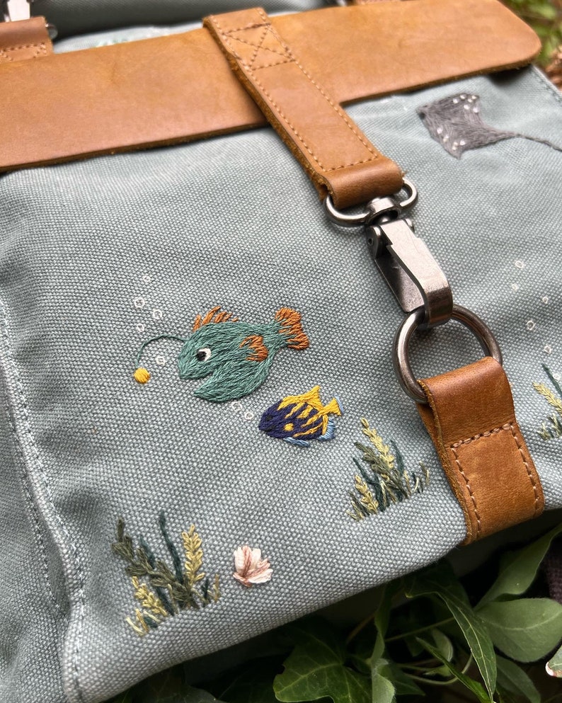 May include: A light blue canvas backpack with a brown leather strap and buckle. The backpack features embroidered images of fish and seaweed.