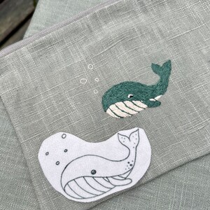 May include: A green fabric pouch with two embroidered whale designs. One whale is outlined in white and the other is filled in with green thread.