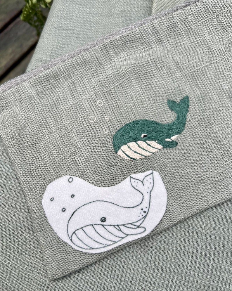 May include: A green fabric pouch with two embroidered whale designs. One whale is outlined in white and the other is filled in with green thread.