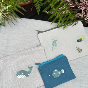 May include: Three small zippered pouches with embroidered sea creatures. The pouches are made of a light gray fabric and a dark blue fabric. The embroidered designs include a whale, a jellyfish, and a fish.