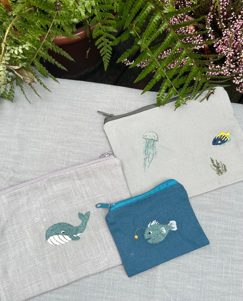 May include: Three small zippered pouches with embroidered sea creatures. The pouches are made of a light gray fabric and a dark blue fabric. The embroidered designs include a whale, a jellyfish, and a fish.