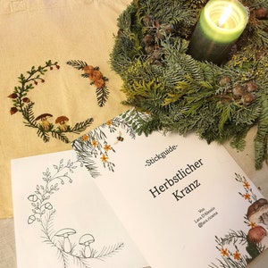 Puede incluir: Un bolso tote beige con un diseño de corona bordada, una corona verde con una vela encendida y una guía titulada "Herbstlicher Kranz". La guía presenta ilustraciones de plantas y setas.