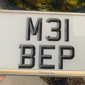 Può includere: Targa rettangolare con la scritta "M31 BEP" in caratteri neri stilizzati. La targa ha una cornice color oro e il marchio "TOYOTA" in basso. Lo sfondo è di colore crema chiaro.