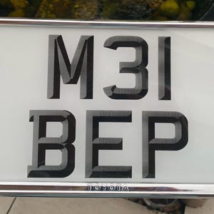 Può includere: Targa rettangolare con la scritta "M31 BEP" in nero su sfondo bianco. La targa ha un bordo argentato lucido e il marchio "TOYOTA" è stampato in basso.
