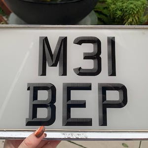 Può includere: Targa rettangolare con i caratteri neri "M31 BEP" su sfondo bianco. La targa è incorniciata in una cornice di metallo argentato lucido. La parola "TOYOTA" è in basso.