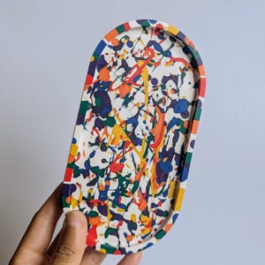 May include: An oval decorative tray with a white base, featuring colourful paint splatters in blue, orange, yellow, and green. The tray's rim has a rainbow border. The tray is being held by a hand.