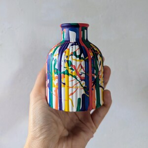 May include: A small, cylindrical vase with a ribbed texture, featuring a vibrant, multi-colored paint drip pattern. The design includes vertical streaks of red, blue, yellow, green, and orange paint over a white base, creating a bold, artistic look.