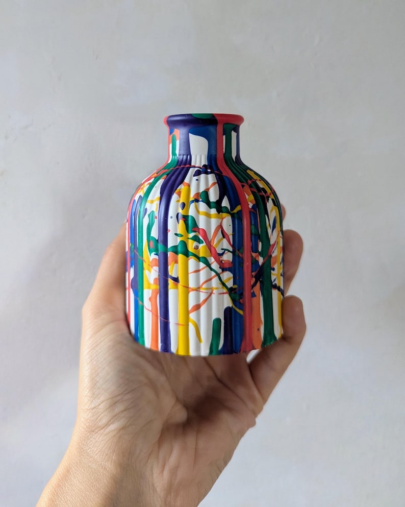 May include: A small, cylindrical vase with a ribbed texture, featuring a vibrant, multi-colored paint drip pattern. The design includes vertical streaks of red, blue, yellow, green, and orange paint over a white base, creating a bold, artistic look.