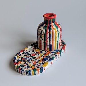 May include: A decorative ceramic vase and matching tray. The vase is white with vertical stripes of red, green, yellow, orange, and blue. The tray is white with colourful paint splatters. The vase has a red rim.