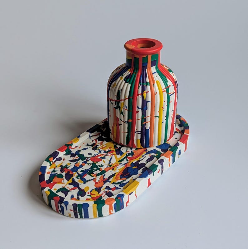 May include: A decorative ceramic vase and matching tray. The vase is white with vertical stripes of red, green, yellow, orange, and blue. The tray is white with colourful paint splatters. The vase has a red rim.