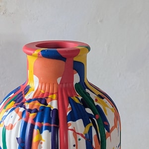 May include: A cylindrical vase with a ribbed texture, featuring a white base color covered in a vibrant, multi-colored paint drip pattern. The design includes streaks and splatters of red, blue, yellow, orange, purple, and green paint.