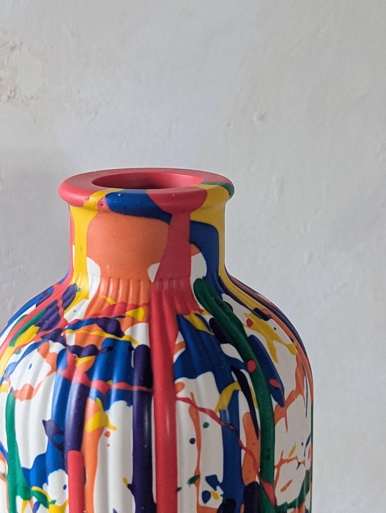 May include: A cylindrical vase with a ribbed texture, featuring a white base color covered in a vibrant, multi-colored paint drip pattern. The design includes streaks and splatters of red, blue, yellow, orange, purple, and green paint.