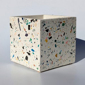May include: A square, off-white planter with a terrazzo design. The planter is speckled with colorful chips of black, blue, green, yellow, pink, and purple. The planter is made of a solid material and has a simple, modern design.